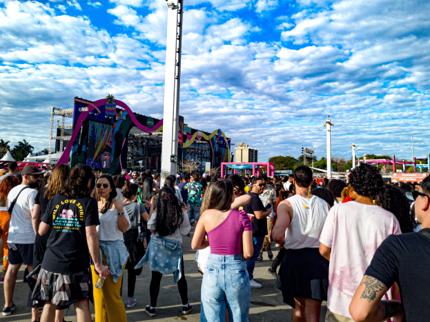Eine Menschenmenge, einige mit Mützen und Schutzbrillen, steht vor einem Fasching mit Stangen, Bäumen, Gebäuden und einem bewölkten Himmel im Hintergrund.