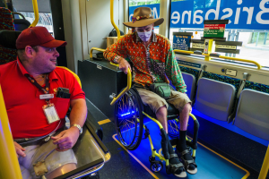 Zwei Männer im Rollstuhl auf einem Bus, einer trägt ein rotes T-Shirt und eine Kappe, der andere ein orangefarbenes Hemd und eine braune Mütze, beide tragen Masken und Ausweise, mit gelben Stangen und Glasfenstern, durch die Bäume, Gebäude und Himmel zu sehen sind.