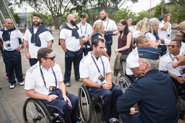 Eine Gruppe von Menschen in Rollstühlen, die vor einer Kulisse aus Bäumen, Pfählen, Gebäuden und einem klaren blauen Himmel miteinander sprechen, wobei einige Mützen und Brillen tragen, was darauf hindeutet, dass sie an den Paralympischen Spielen teilnehmen.