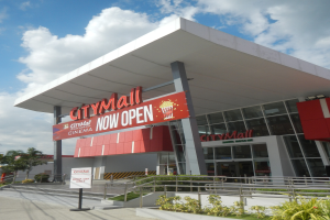 Ein belebter Stadtplatz mit einem Schild "City Mall Now Open" umgeben von Gebäuden, Namensschildern, Geländern, Pflanzen, Bäumen, Strommasten mit Drähten und einem bewölkten Himmel im Hintergrund.