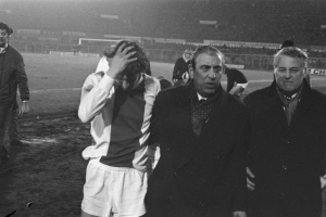 Eine Gruppe von Männern in Not steht zusammen auf einem Fußballfeld mit einem Stadion im Hintergrund, schwarz-weiß.