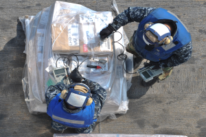 Zwei Männer in blauen Uniformen und Helmen arbeiten an einem großen Drohnenmodell und halten verschiedene Gegenstände in den Händen, mit einer Plastikabdeckung vor ihnen auf dem Boden.