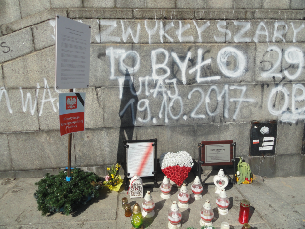 Ein Denkmal mit Blumenstr"{a}uen, Kerzen und anderen Gegenst"{a}nden vor einer steinernen Wand mit Text und Graffiti, wahrscheinlich im Gedenken an Holocaust-Opfer.