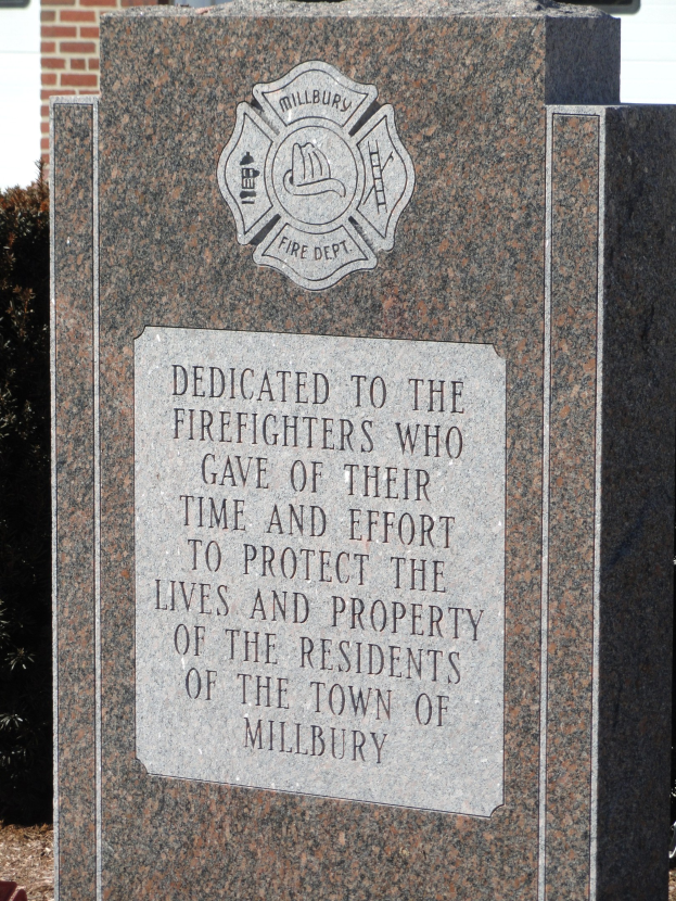 Gedenkstein mit einer Plakette, die Feuerwehrleuten in Millbury gewidmet ist, vor einem Backsteingebäude mit umliegender Vegetation.