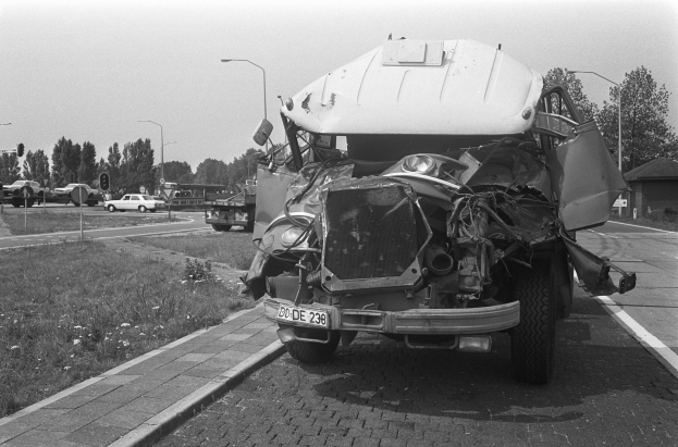 Ein Schwarz-Weiß-Foto eines schwer beschädigten Autos am Straßenrand, dessen Motorhaube eingedellt und Fenster zersplittert sind, während andere Fahrzeuge, Laternenpfähle, Bäume und ein Haus im Hintergrund zu sehen sind.