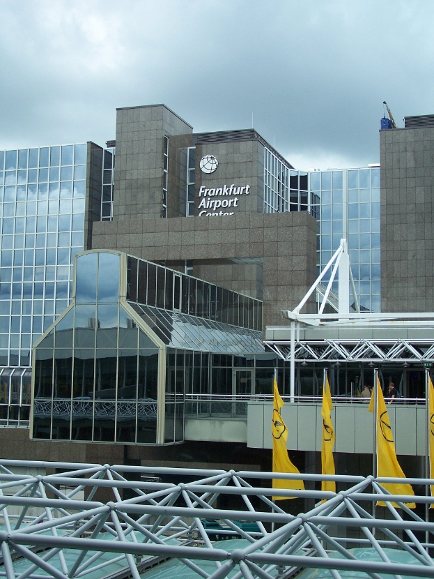 Frankfurt Airport in Frankfurt, Germany, ein großes Gebäude mit Glaswänden und Text, umgeben von gelben Flaggen und Eisenstangen, unter einem Himmel voller weißer, flauschiger Wolken.