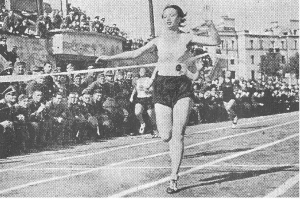 Ein Schwarz-Weiß-Foto einer Frau, die auf einer Laufbahn während des Frauen-100m-Finals bei den Olympischen Spielen 1956 läuft, mit einer Zuschauermenge, Gebäuden und Pfosten im Hintergrund.