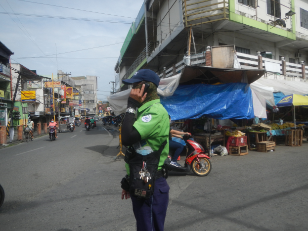 Ein Polizeibeamter in grüner und blauer Uniform und blauer Möppe steht an der Straßenseite und spricht in ein Handy, mit Fahrzeugen im Hintergrund, Ständen mit Gegenständen rechts von ihm und einer bewölkten Himmel über ihm.