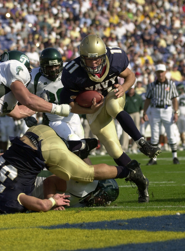 Ein Fussballspieler in einem Sports Outfit rennt mit dem Ball, umgeben von anderen Spielern mit Helmen auf einem Feld, mit einem Schiedsrichter und einer Person in einer M├╝tze im Hintergrund, das Bild ist leicht unscharf.