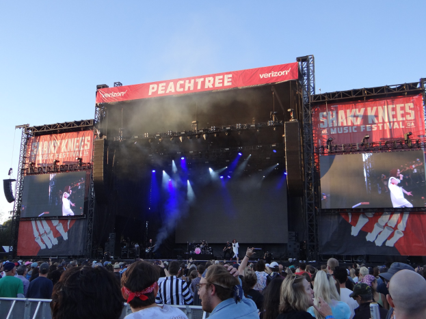 Eine große Menschenmenge vor einer hell erleuchteten Bühne beim Shaky Knees Music Festival, wo Musiker hinter Bannern spielen, mit einem Geländer im Vordergrund und einem klaren blauen Himmel im Hintergrund.