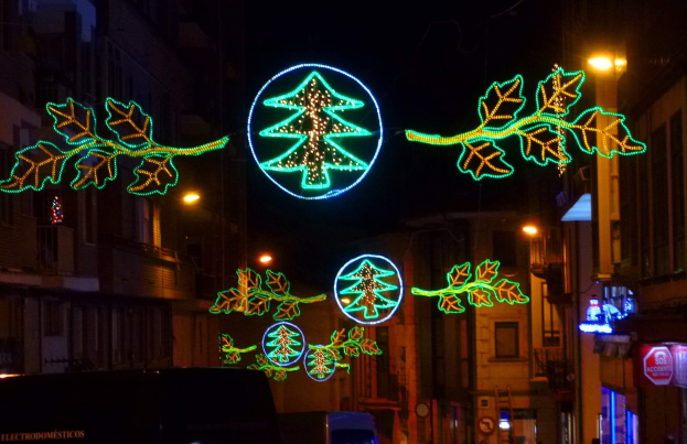 Eine nächtliche Stadtstraße, dekoriert mit festlichen Weihnachtslichtern in Form von Blättern, mit Fahrzeugen auf der Straße und Gebäuden auf beiden Seiten.