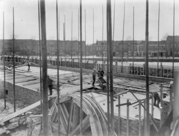 Schwarz-weiß-Foto einer Baustelle mit Arbeitern, Holzpfählen und einem Rad im Vordergrund, Gebäuden, Bäumen und einem klaren Himmel im Hintergrund.