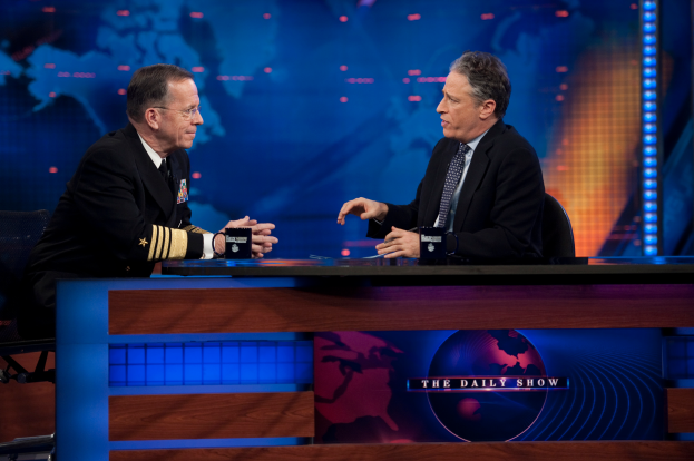 Zwei Männer in schwarzen Blazern, Hemden und Krawatten sitzen an einem Tisch mit Mikrofonen zwischen ihnen und führen ein Gespräch vor einem TV-Bildschirm und einem blauen Hintergrund.