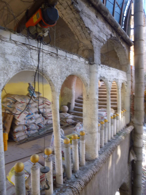 Altes Gebäude mit bögen Fenstern, Säulen und verstreuten Werkzeugen und Materialien, darunter eine Maschine oben und Gegenstände am Boden.