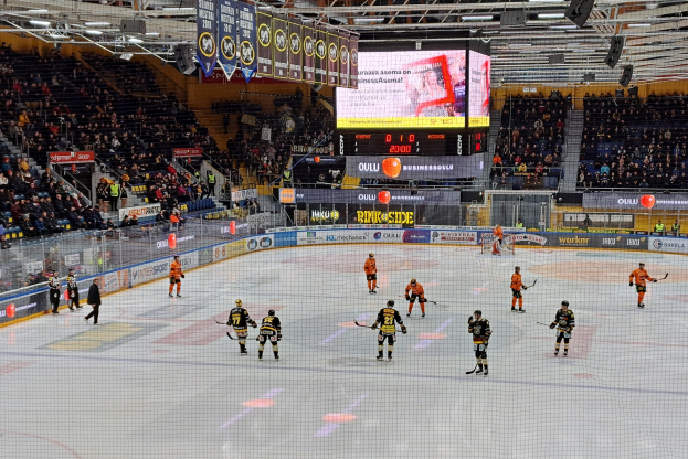 Eine Gruppe von Menschen mit Helmen und Hockeystöcken spielt ein Spiel auf einem Eisstadion mit einem Netz in der Mitte, umgeben von Texttafeln und einem großen Bildschirm im Hintergrund, mit einer Person mit Hut im Bild.