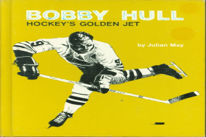Bobby Hull, ein Eishockeyspieler, hält einen Eishockeyschläger, trägt einen Handschuh und zeigt ein Buch mit dem Titel "Hockeys Golden Jet" von Julian May.
