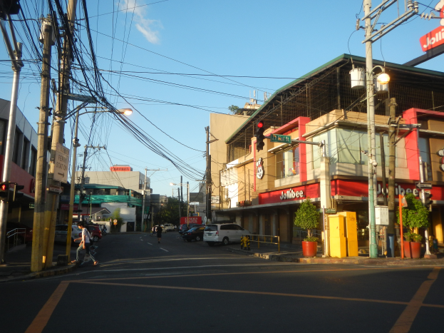 Eine belebte Stadtstraße mit Fußgängern, Fahrzeugen und verschiedenen städtischen Elementen wie Verkehrszeichen, Gebäuden und Pflanzen unter einem bewölkten Himmel.