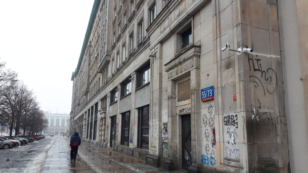 Eine Person geht auf einem Gehweg an einem Gebäude mit Graffiti vorbei, mit parkenden Fahrzeugen auf der Straße, Bäumen auf beiden Seiten und Gebäuden im Hintergrund unter dem Himmel.