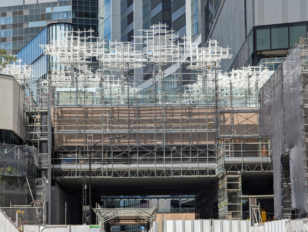 Groes Bauwerk mit Geruest, umgeben von Baumen links und einer Strasse unten in einer stadtischen Umgebung.