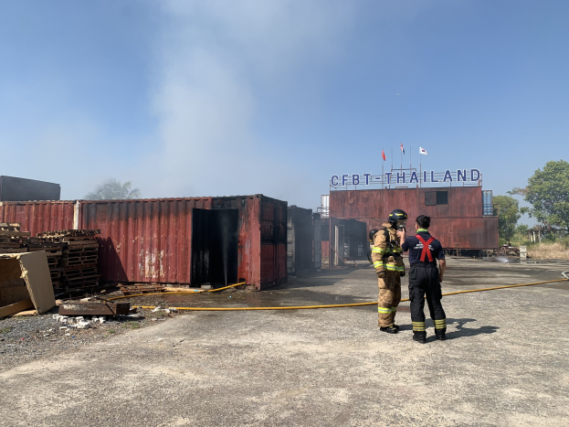 Zwei Feuerwehrleute in Helmen vor einem rauchenden Gebäude, einer hält ein Rohr, mit verstreuten Trümmern und einem Schild in der Nähe, vor einem Hintergrund aus Bäumen und einem blauen Himmel.