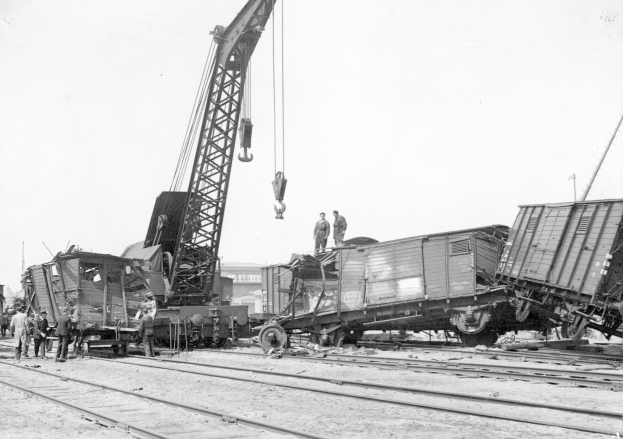 Schwarß-weißes Bild eines Zugunglücks mit einem Kran, der ein Zugwaggon auf die Schienen hievt, mehrere Menschen in der Nähe, Gebäude und ein klarer Himmel im Hintergrund.