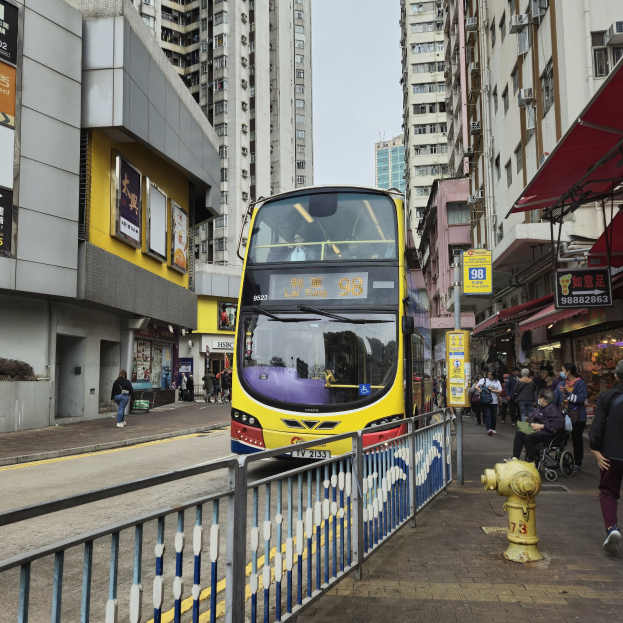 Ein gelber Doppeldeckerbus fährt eine von hohen Gebäuden gesäumte Straße entlang, mit Menschen auf dem rechten Gehweg, einem Hydranten und einem Geländer, unter einem klaren blauen Himmel.