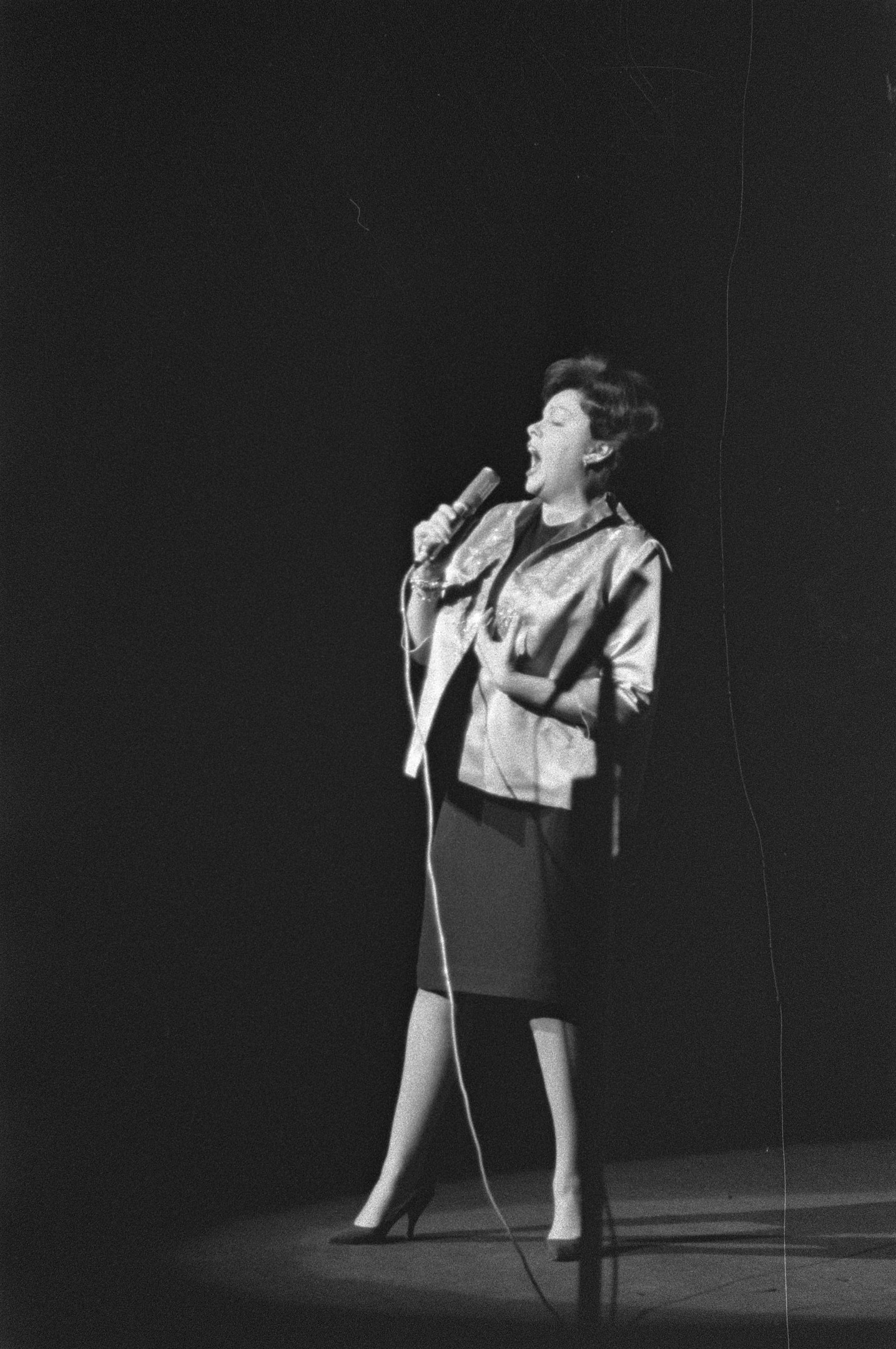 Eine Frau in einem schwarzen und weißen Outfit aus den 1950er-Jahren singt fröhlich auf einer von einem Scheinwerfer beleuchteten Bühne und hält ein Mikrofon.