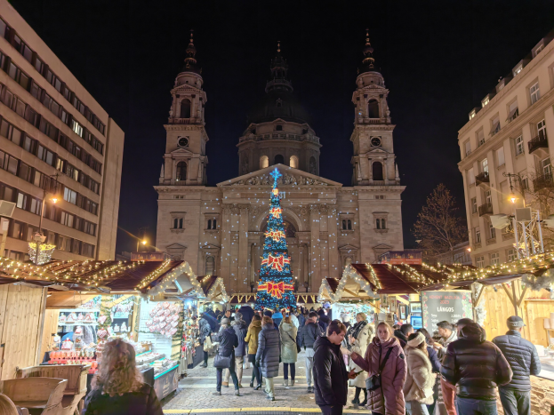 Ein belebter Weihnachtsmarkt mit festlich geschmückten Ständen und Lichtern vor einer Kirche bei Nacht, umgeben von Gebäuden, Bäumen und einem sternenklaren Himmel.