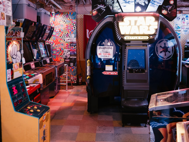 Ein hell erleuchteter Arcade-Raum mit mehreren Maschinen, darunter ein Star Wars Battle Pod, farbige Poster an den Wänden und verstreute Gegenstände, die eine lebendige Atmosphäre schaffen.