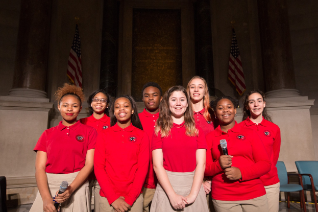 Eine Gruppe junger Frauen in roten Hemden, die strahlen und Mikrofone halten, vor Stühlen, Säulen und Fahnen stehen, wahrscheinlich Teil eines nationalen Jugendchors.