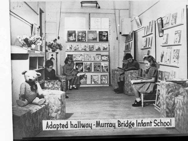 Gruppe von Kindern auf Stühlen in einem bibliotheksähnlichen Raum mit Bücherregalen, einem Teddybär links, einer Vase auf dem Tisch, gerahmten Gegenständen an der Wand und Text unten mit "Angepasster Flur Murray Bridge Kindergarten"
