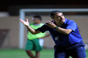 Ein Mann in einem blauen T-Shirt und Hose steht auf einem Fußballfeld und hält ein Mikrofon in der Hand, während er auf seine Spieler zeigt. Im Hintergrund sind eine Person in einem grünen Kleid, ein Tor und eine Wand zu sehen.