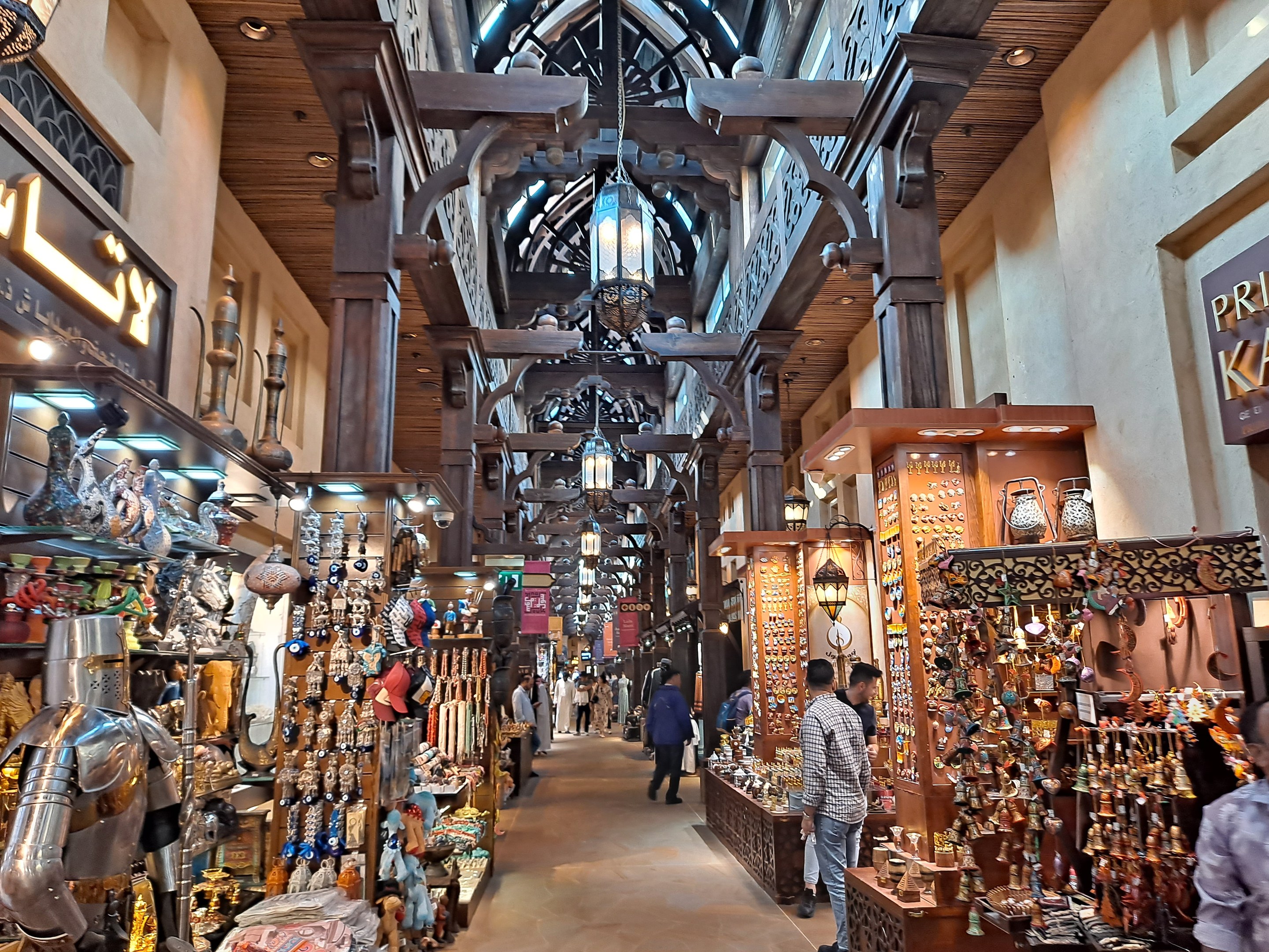 Ein belebter Dubai-Basar mit Menschen, die zwischen Regalen voller Waren gehen, beleuchtet von Deckenlampen und mit Texttafeln an den Wänden.