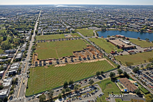 Luftaufnahme einer Stadt mit Gebäuden, Bäumen, Gras, Straßen mit Fahrzeugen, Wasser und Wolken, die ein großes zentrales Feld zeigen, das für einen Park vorgesehen ist.
