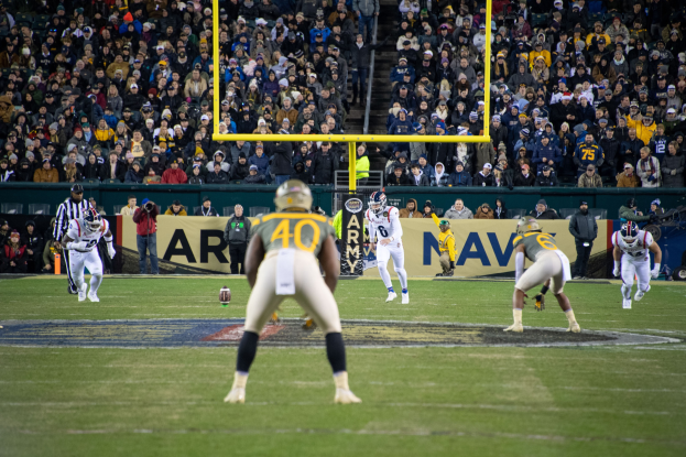 Fußballspieler in Sportkleidung und Helmen konkurrieren auf einem Stadionfeld mit Zuschauern im Hintergrund und Beschriftungen auf Pfählen und Tafeln.