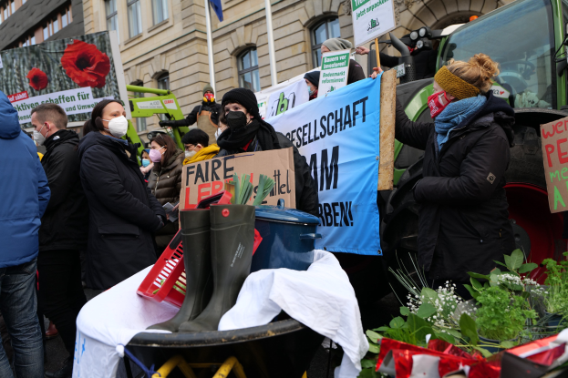 Eine Gruppe von Menschen in Masken mit Protestschildern vor einem Lastwagen, mit einem Tisch, Pflanzen, Gebäuden und einer Flagge im Hintergrund.