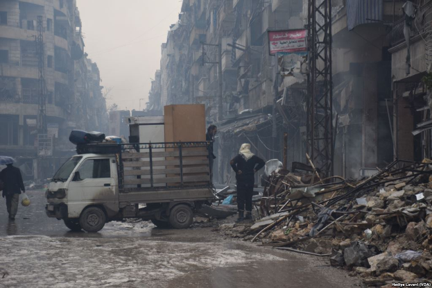 Ein Lastwagen fährt auf einer Straße neben einem Schutthaufen, mit Menschen, die auf der Straße gehen, einer hält einen Schirm, und Gebäuden, Polen und Brettern mit Text im Hintergrund unter einem klaren Himmel.