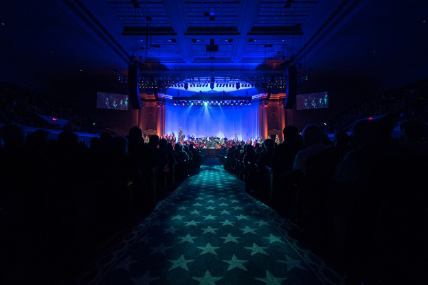 Ein großer Auditorium mit in Reihen sitzenden Menschen, eine Bühne mit Musikern und Deckenlampen und Fernseher, was auf eine Konzerthalle hinweist.