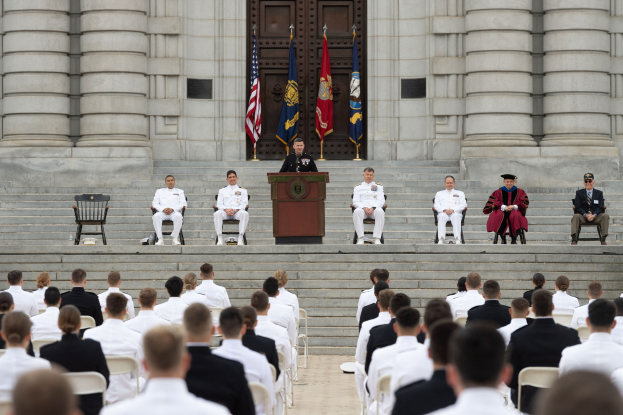 Gruppe von Menschen, die auf Stühlen sitzen und auf ein Gebäude mit Säulen und einer Tür schauen, mit einem Podium und einem Mikrofon in der Mitte, Fahnen im Hintergrund und einem leeren Stuhl links.