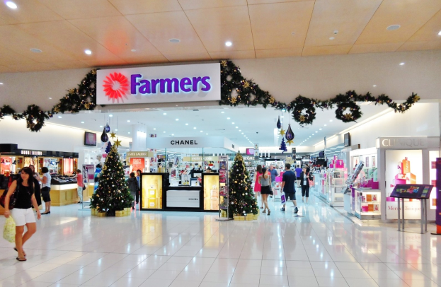 Eine Gruppe von Menschen, die durch einen weihnachtsgeschmückten Einkaufszentrum geht, mit beleuchteten Bäumen, Dekorationen und einer Deckenanzeige, einige tragen Einkaufstaschen.