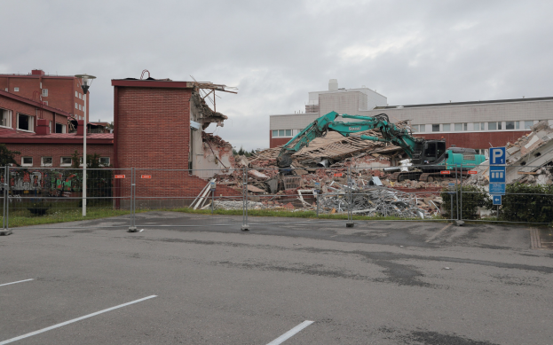 Groes Bauwerk in einer Parkplaetze mit einem Bagger, Metallstangen, einem Zaun, einem Schild, Pflanzen, Gras, Baeumen, naeheren Gebaeuden und einem bewolktem Himmel.