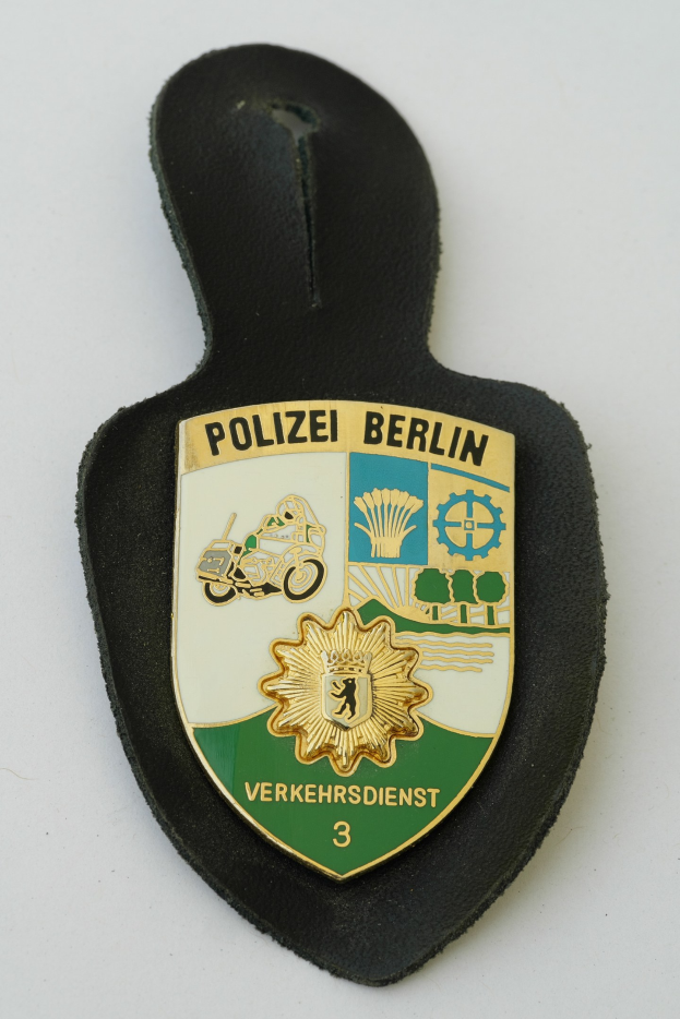 Ein schildförmiges Polizeiamblem mit der Aufschrift 'Polizei Berlin' auf einem blauen Hintergrund, einem weißen Rand, einem weißen Stern in der Mitte und schwarzer fetter Schrift, das auf einer Oberfläche liegt.