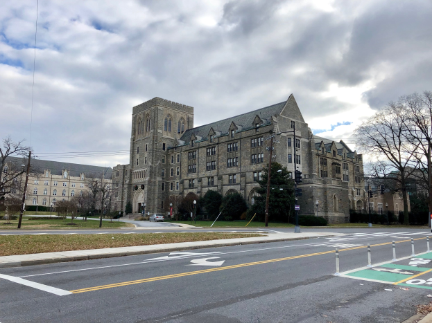 Großes Gebäude mit vielen Fenstern, als Universität von Pennsylvania beschriftet, umgeben von Bäumen, Straßenlaternen, Verkehrssignalen und Fahrzeugen an einem bewölkten Tag.