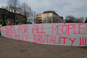Eine Gruppe von Menschen hält ein Transparent mit der Aufschrift 'Rechte für alle Menschen Stoppt Polizeigewalt' auf dem Boden mit einem Straßenschild, einem Schild, Bäumen, Gebäuden mit Fenstern und einem bewölkten Himmel im Hintergrund.