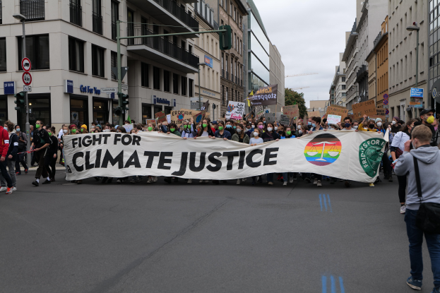 Eine Gruppe von Menschen marschiert mit einem "Kampf für Klimagerechtigkeit"-Schild die Straße entlang, mit Gebäuden, Schildern, Ampeln und Bäumen am Straßenrand unter einem bewölkten Himmel.