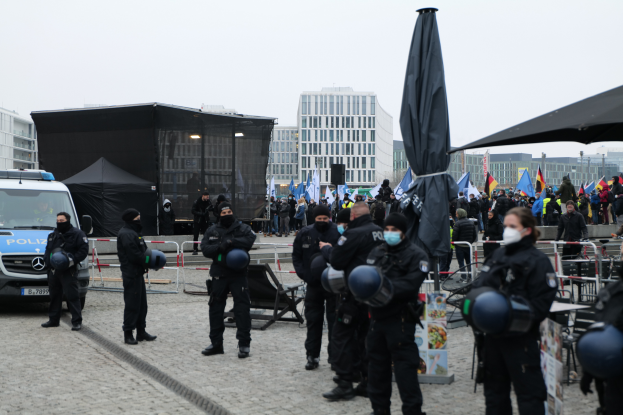 Eine Gruppe von Polizisten steht vor einer Menge von Demonstranten in Berlin, Deutschland, mit Barrikaden, Zelten und Fahnen im Vordergrund und Gebäuden unter einem klaren Himmel im Hintergrund.