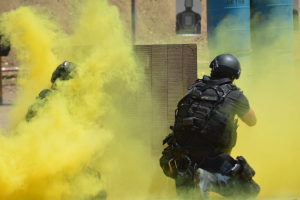 Zwei Polizisten in Einsatzausrüstung mit gelbem Rauch aus dem Mund, vor einer Wand mit einer Tafel und anderen Gegenständen und dem Boden unten.