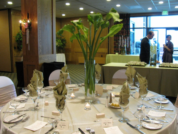 Ein Hotelesszimmer mit einem weißgedeckten Tisch, auf dem Teller, Besteck, ein Glas und Zweige liegen, umgeben von Stühlen, einem grünen Tisch im Hintergrund, einem Mann in einer schwarzen Jacke, einem stehenden Mädchen, einer Tür mit der Aufschrift "Ausgang" und zwei Männern dahinter.