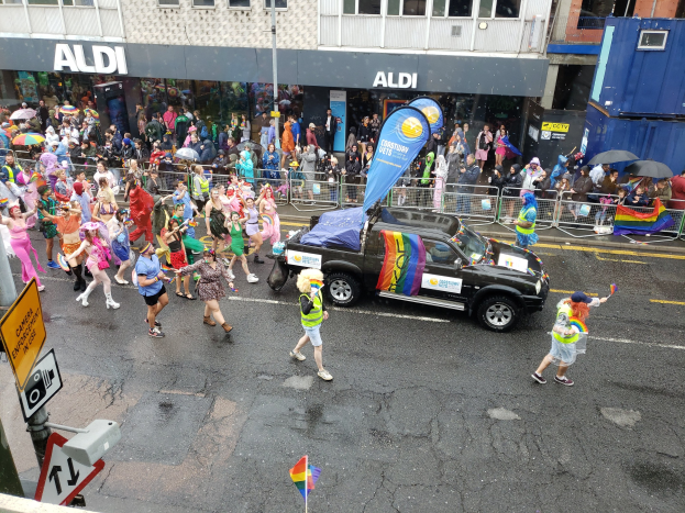Eine Gruppe bunt gekleideter Menschen, die auf einer Straße in einer Parade marschieren, mit einem Auto in der Mitte, einige halten Schirme, und Gebäude mit Fenstern im Hintergrund.