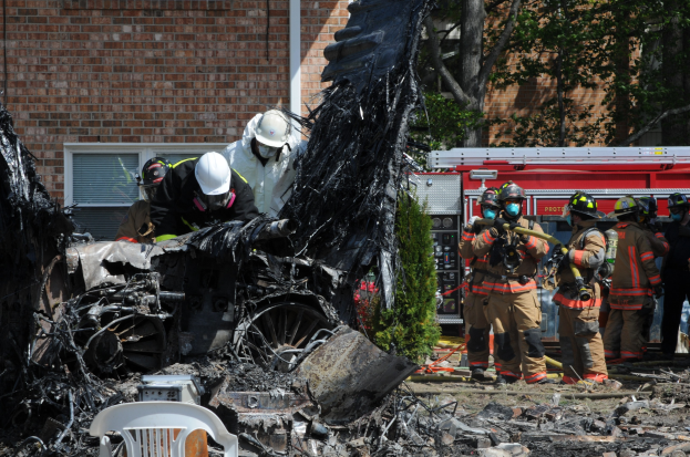 Feuerwehrleute in Schutzausrüstung löschen ein Gebäude Feuer, mit verkohlten Überresten einschließlich eines Fahrzeugs, Stuhls und Pflanzen auf dem Boden.